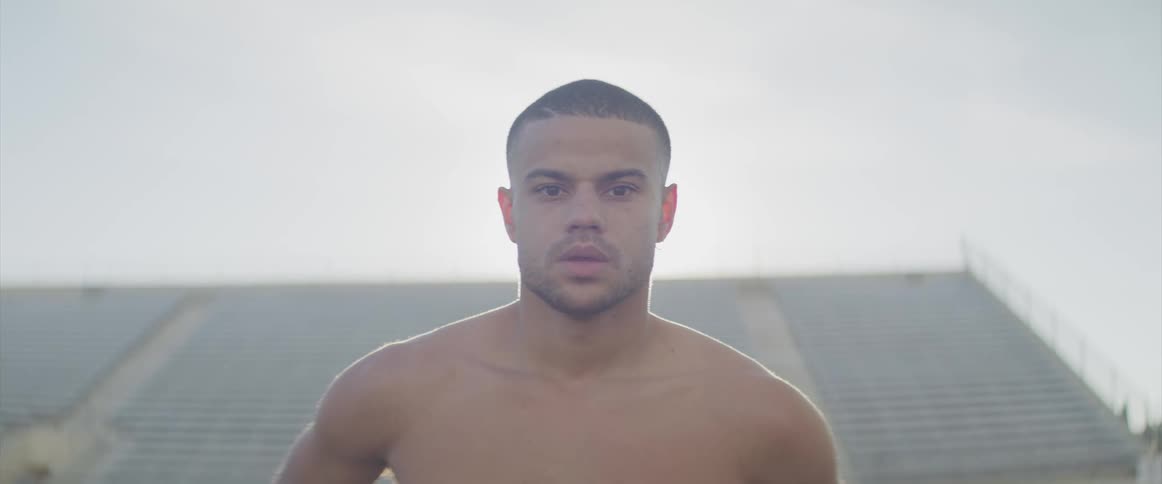 A shirtless young man takes deep breaths as he looks into the camera while standing on an athletics field on a sunny day