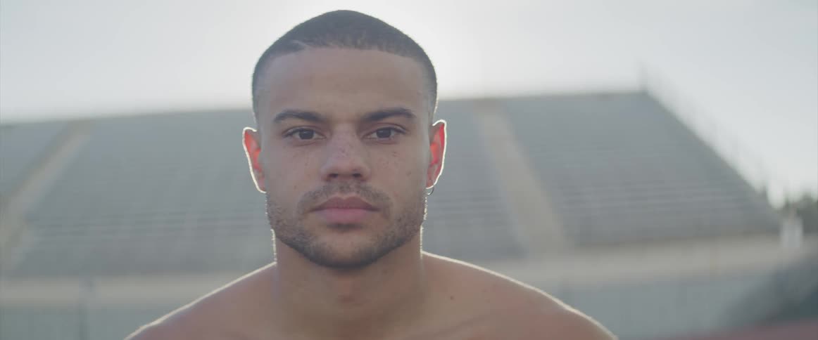 A young shirtless male stands before seating stands on a very sunny day