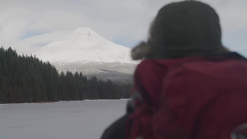 A person in a red coat stands outside in the snow and looks toward a snow covered mountain peak