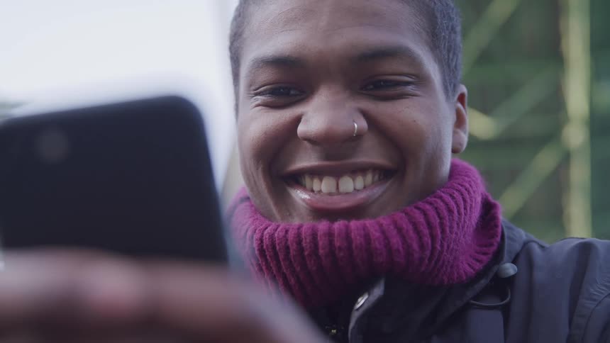 Male wearing a nose ring and heavy sweating sits outdoors while smiling and texting on his smart phone