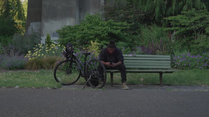 A man with a smart phone sitting on a bench next to a bike
