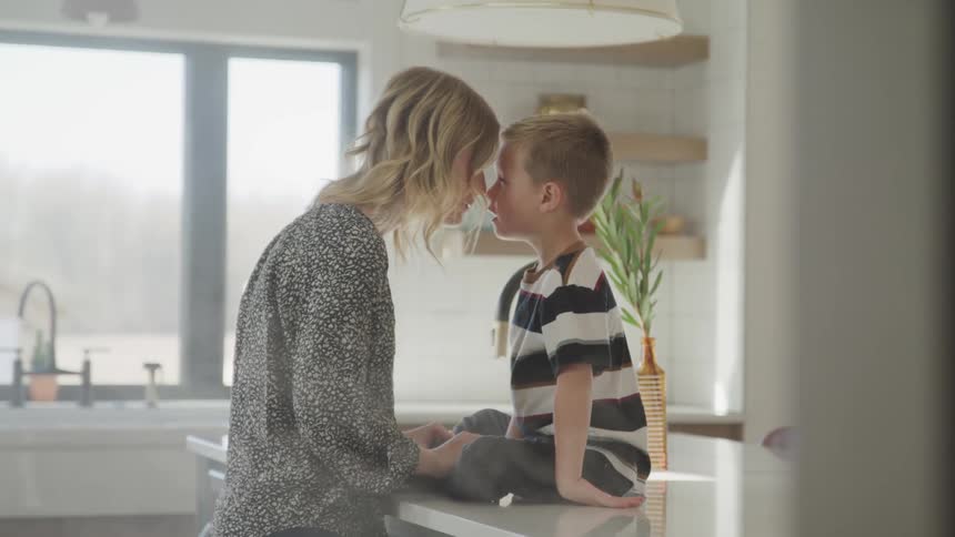Mother rubs nose against sons nose as he sits on kitchen island counter