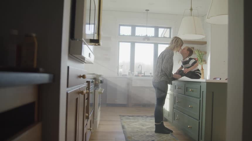 Young boy sits on a kitchen counter as his mother stands in front of him and kisses him