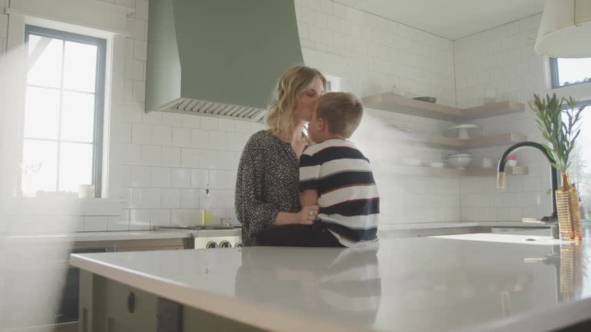 A little boy sits on the kitchen island and his mom leans forward and kisses his forehead