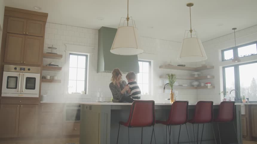 A little boy sits on the kitchen island and his mom tickles him and he laughs