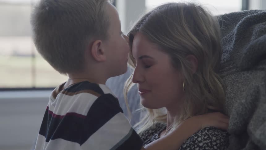 A woman sits with her eyes closed on the floor of a bedroom as a little boy kisses her on the forehead