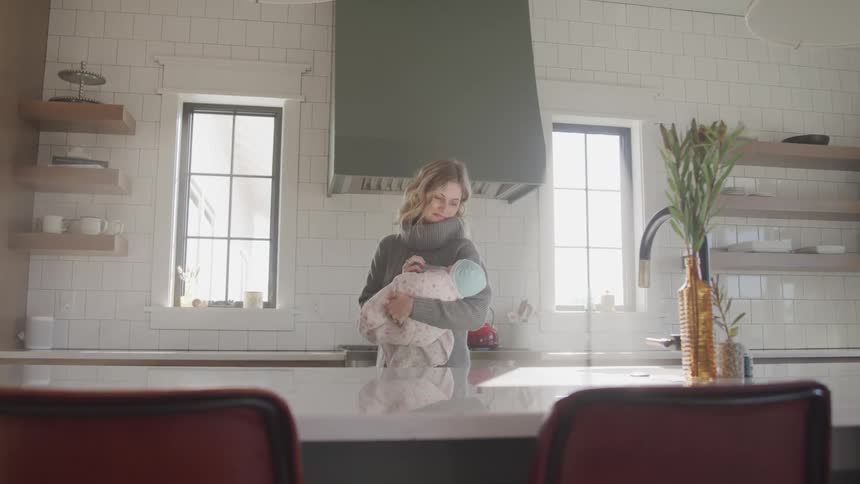 A woman holds an infant in her arms standing in the kitchen