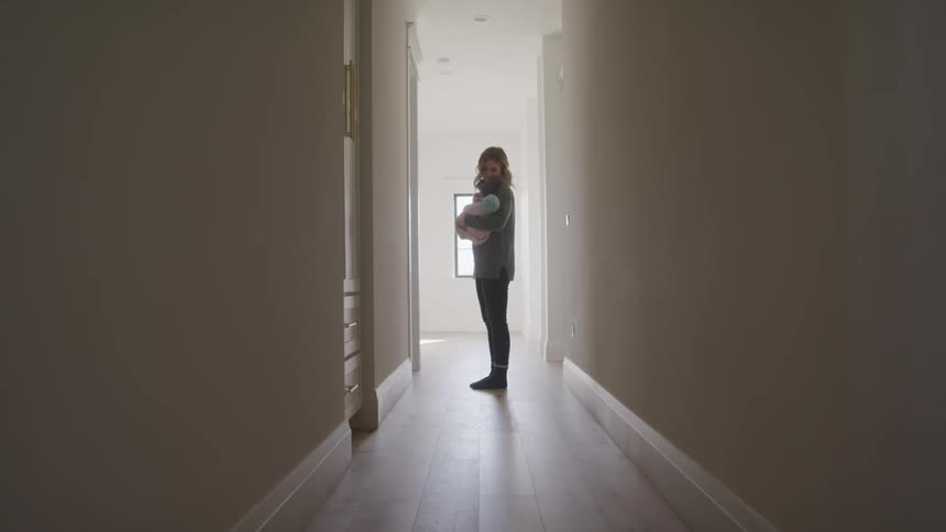 Woman wearing sweater with cowl stands in hallway smiling down at baby in her arms