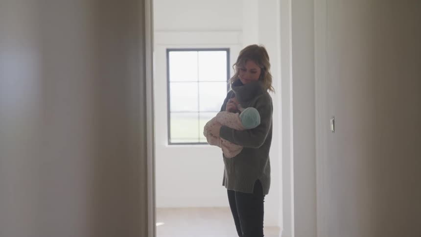 Mother smiles while feeding and holding infant standing in room near hallway