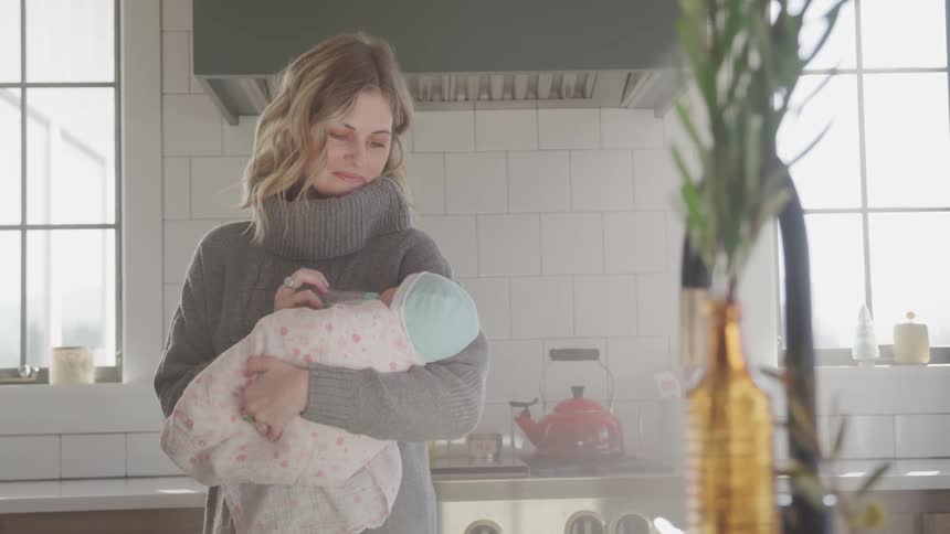 A woman stands in her kitchen and rocks back and forth as she feeds her baby she is holding in her arms