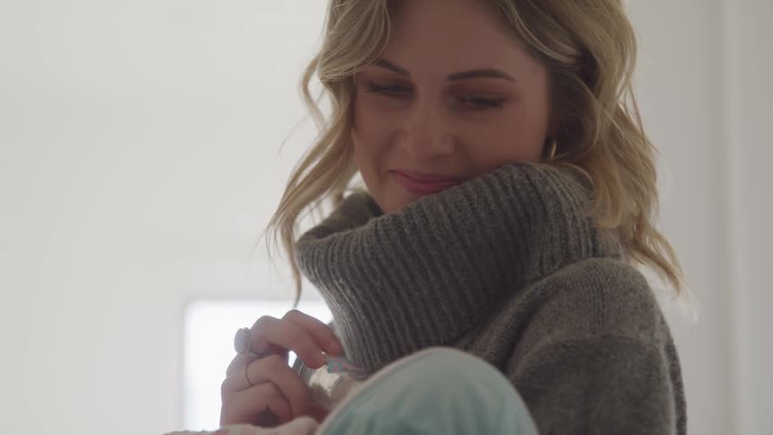 Mother wearing sweater cowl holds baby and bottle shes using in feeding