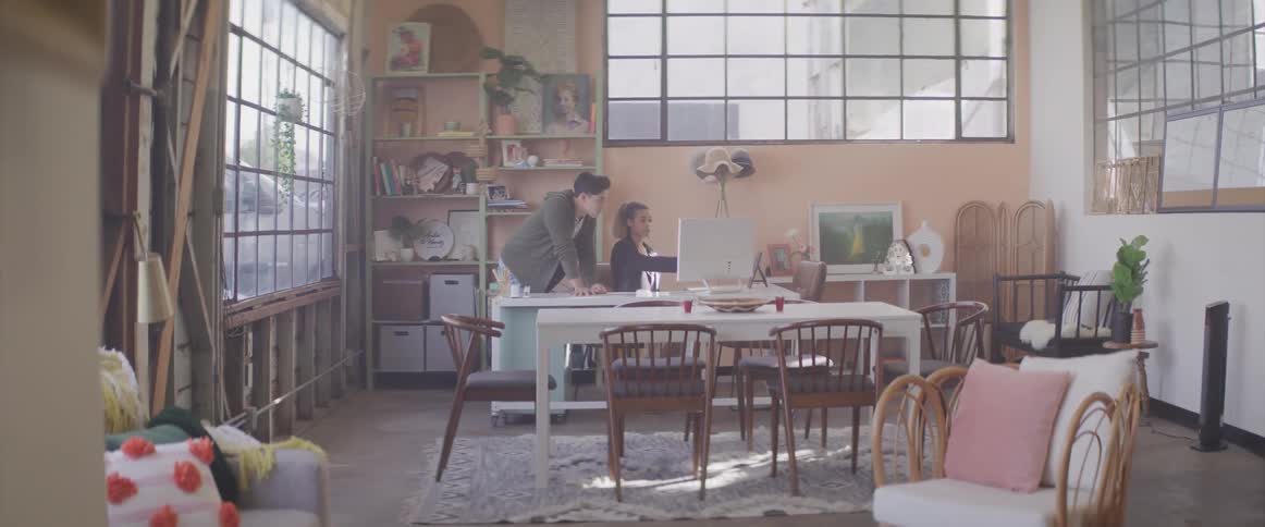 A young woman sitting at a desk points at her computer monitor as a young man standing behind her watches