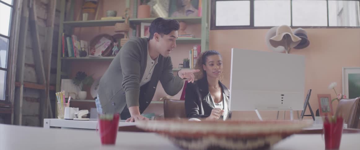 A man and woman sit at a computer desk talking about what they see on the screen