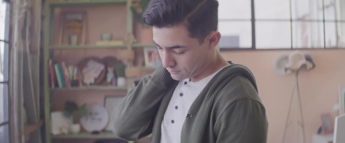 A young man standing indoors looks downward was he places his hand on the back of his neck