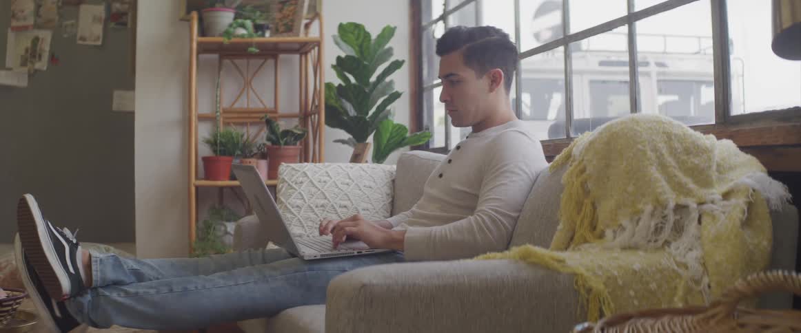 A man sits on his couch with his laptop on his knee and types on it