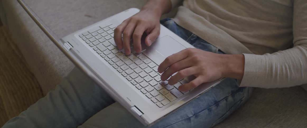 Man with jeans and a brown sweatshirt sits on a couch and types on a grey laptop
