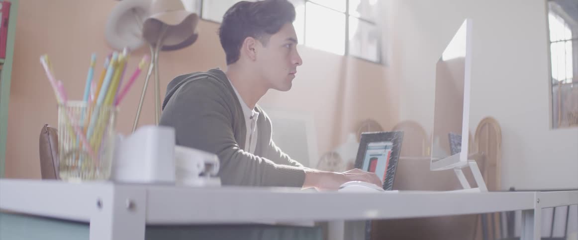 Man wearing hoodie sits typing on computer as sun glares through window