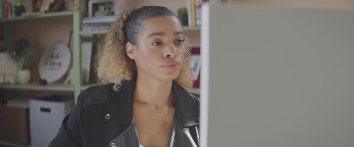 Woman with curly hair and a black jacket sits at a desk and uses a computer