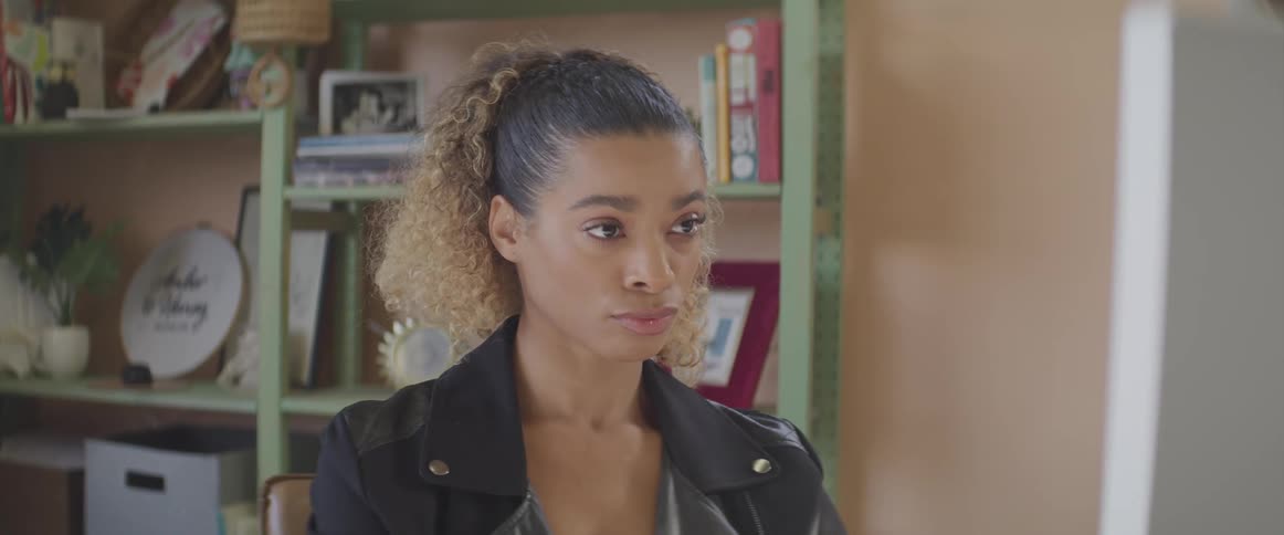 Black woman with curly hair and a leather jacket sits at a desk and uses a computer in a room