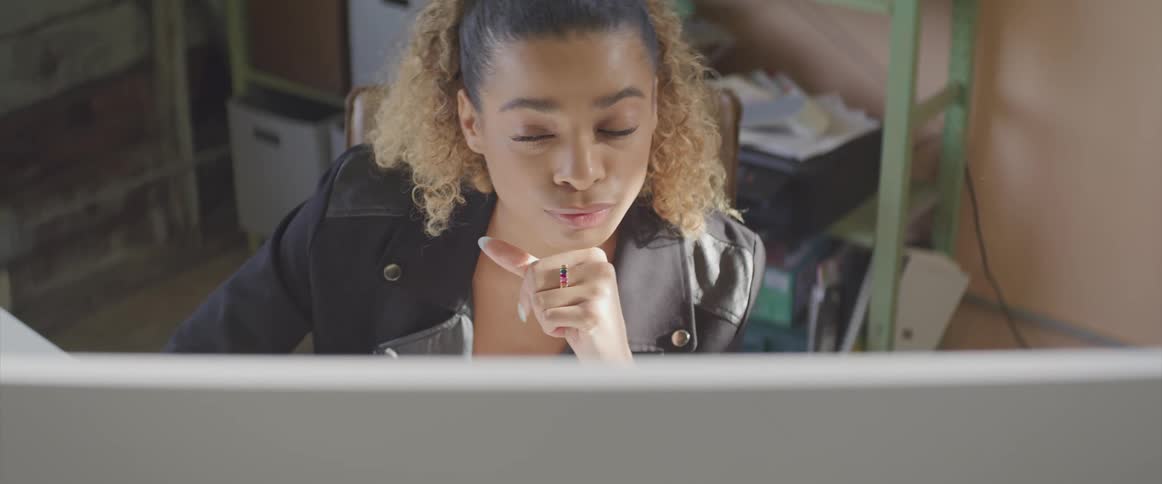 Black woman with curly brown hair and a black leather jacket sits at a desk and uses a computer
