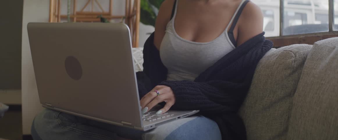 Brunette woman with a grey undershirt and jeans sits on a grey coach and uses a grey laptop in the living room