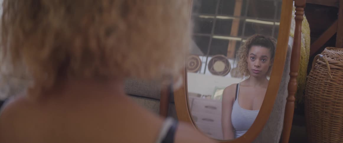 A young woman looks at her reflection in the mirror as she sits on the floor of a living room