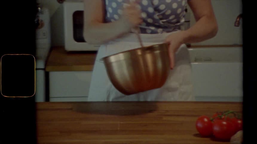 Female wearing an apron in the kitchen uses a whisk to aggressively stir ingredients together in a silver metal bowl