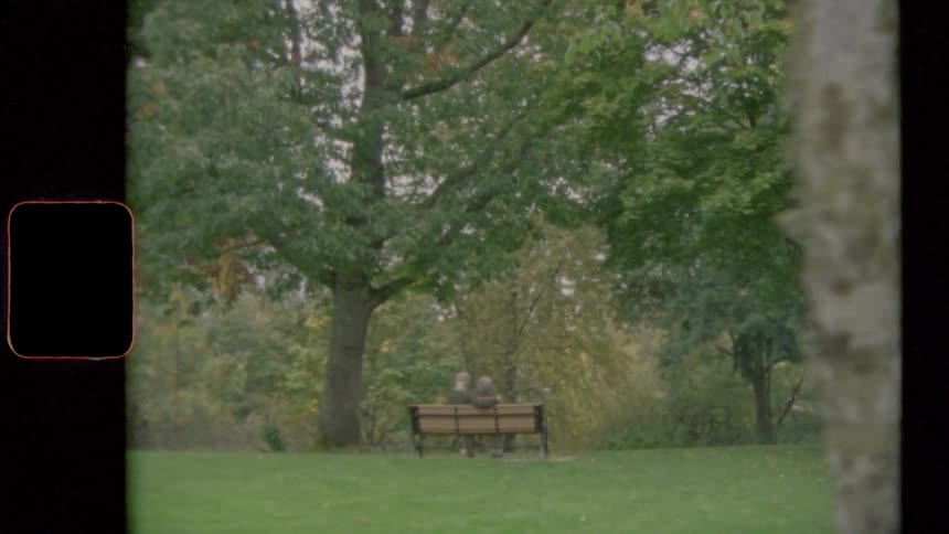 Couple sit on park bench together on edge of mowed grass facing plantings
