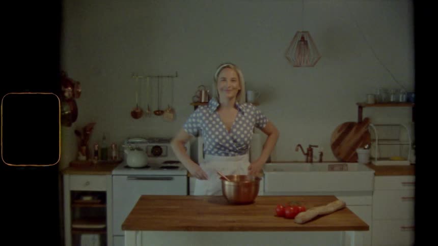 A woman stands in a kitchen stirring the contents of a mixing bowl before posing and smiling