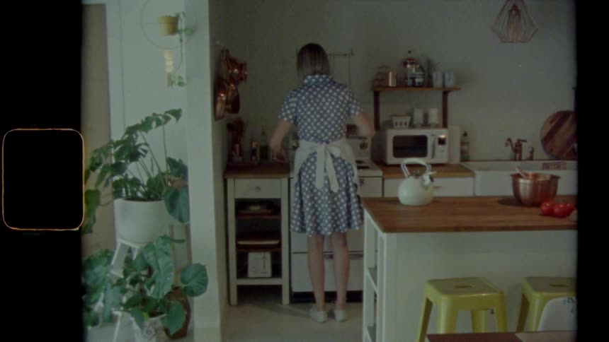 A woman wearing a dress and apron stands in a kitchen while preparing food