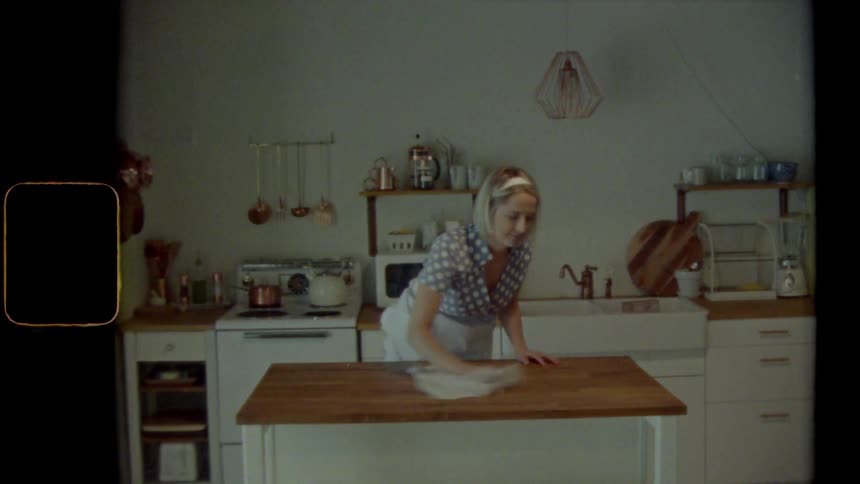 Blonde woman with a blue dress and a white apron cleans a table with a towel in the kitchen