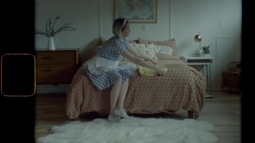 A woman wearing a dress sits on a bed while folding a piece of clothing