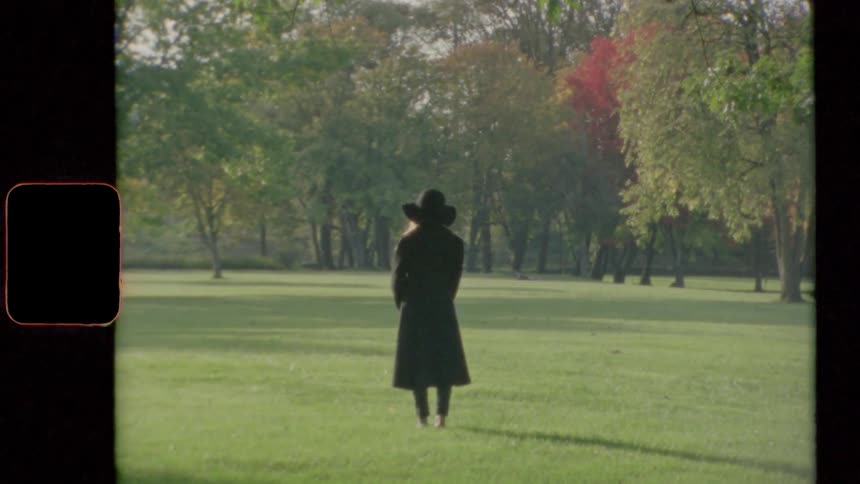 A woman with a coat and a hat standing on grass with trees and the sky in the background