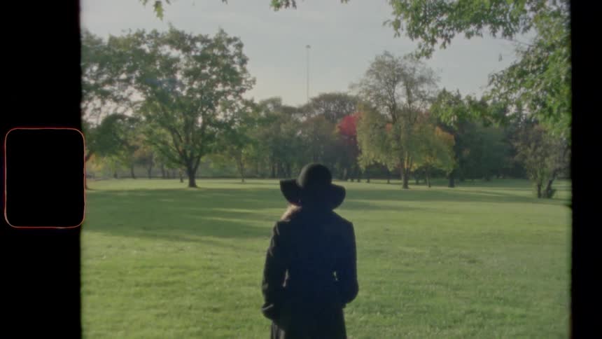 Woman wearing a coat and a brimmed hat stands on the grass looking out at the forest