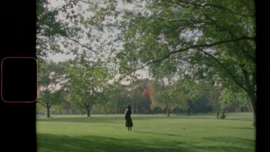 Woman with a black topcoat and a hat stands on green grass in public park