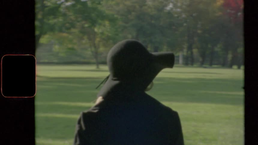 Woman with a black topcoat and a hat stands on green grass in public park