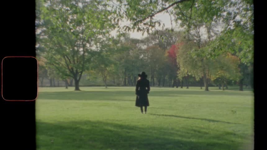 A woman stands in an open field in a park wearing a black trench coat and black felt hat