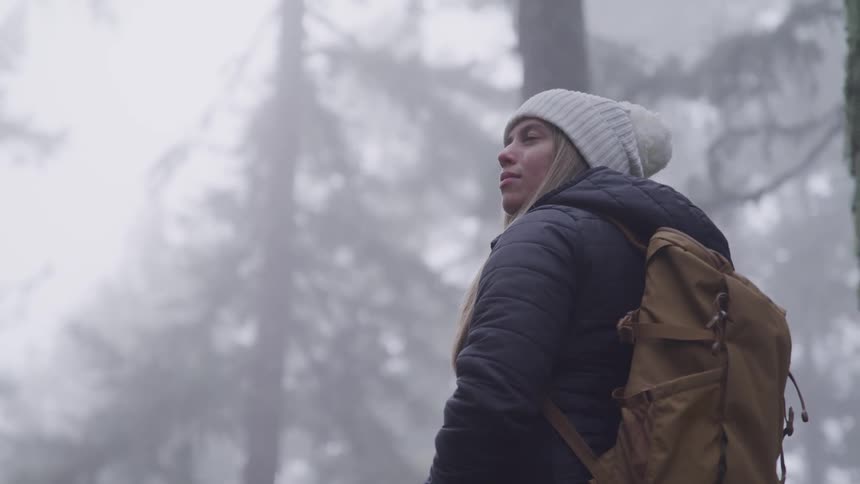 Woman with a with a white beanie a black coat and a brown backpack looks to the right in the foggy forest