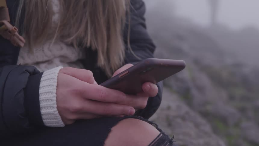 A woman sits on a rock while typing on her smartphone outdoors on a foggy day