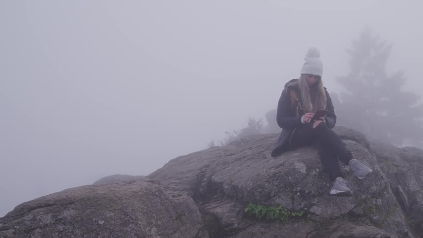 A teenage girl sits on a rock outside in the fog and uses her cell phone