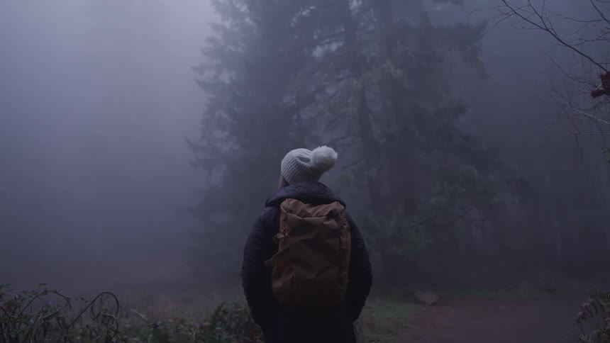 Woman with a white beanie a black coat and a brown backpack stands in a dark foggy forest