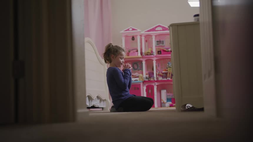 A young girl kneels on her bedroom floor with her hands clasped in prayer