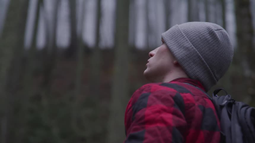 Man with a grey beanie red coat and a black backpack looks up at trees in the forest