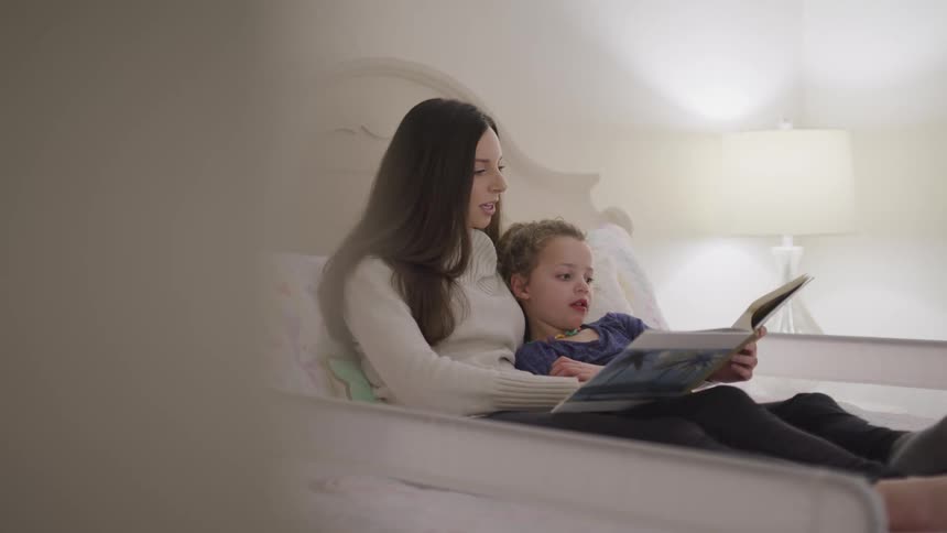 Daughter lying next to mother in a twin size bed listening intently as mother reads daughter a story book by the light of the lamp