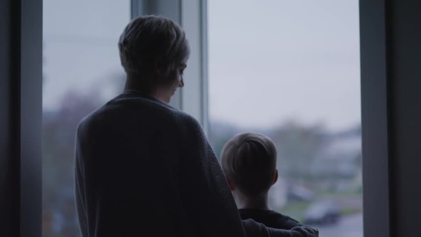 Mother and son looking out the window are silhouetted against the cloudy sky