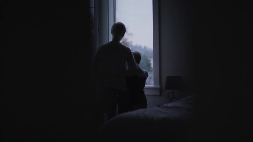 Mother stands near window hugging both children about the shoulders