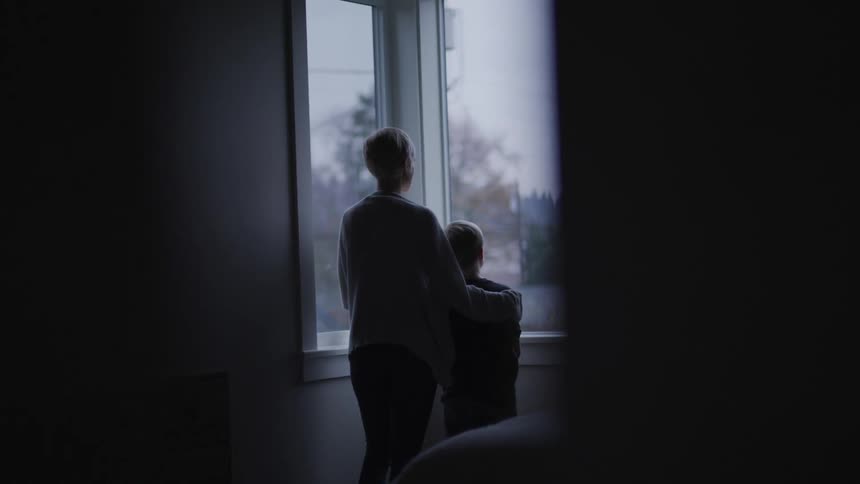 Mother and son looking out of the window of a dimly lit bedroom at home