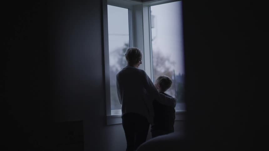 A mother stands with her arm around her son and they look out the window at the snow