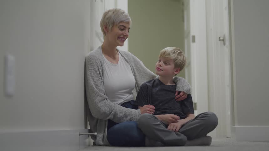 A woman and a boy sitting in a hallway floor with doors in the background