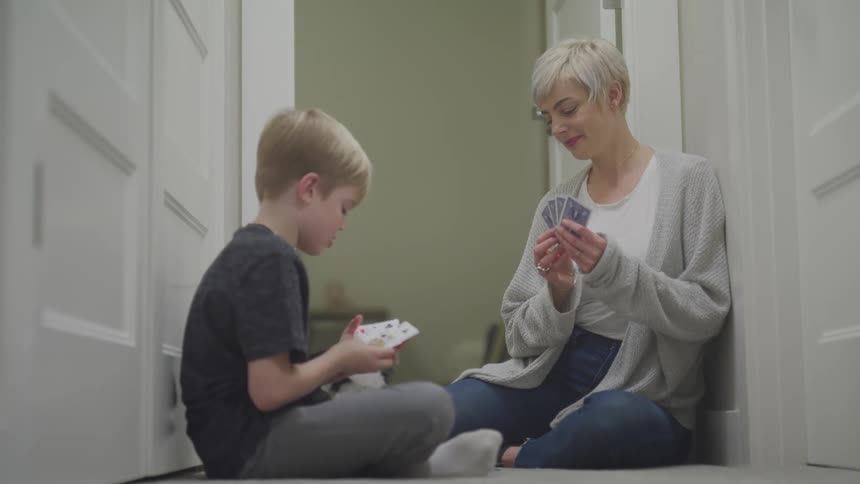 A white woman with short blonde hair and a sweater and jeans sits with a white boy with brown hair and a tshirt with sweatpants sit in a hall playing a card game